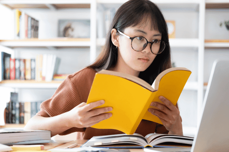 A young woman with glasses reading a yellow book while studying at a desk with a laptop.