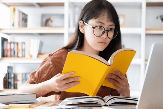 A young woman with glasses reading a yellow book while studying at a desk with a laptop.