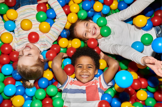 Three smiling children lying in a colorful ball pit, surrounded by red, yellow, green, and blue plastic balls