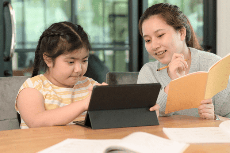 Young girl using a tablet for learning while her mother guides her with a notebook and pencil.