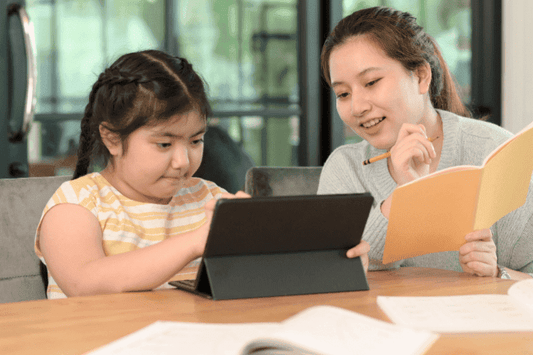 Young girl using a tablet for learning while her mother guides her with a notebook and pencil.