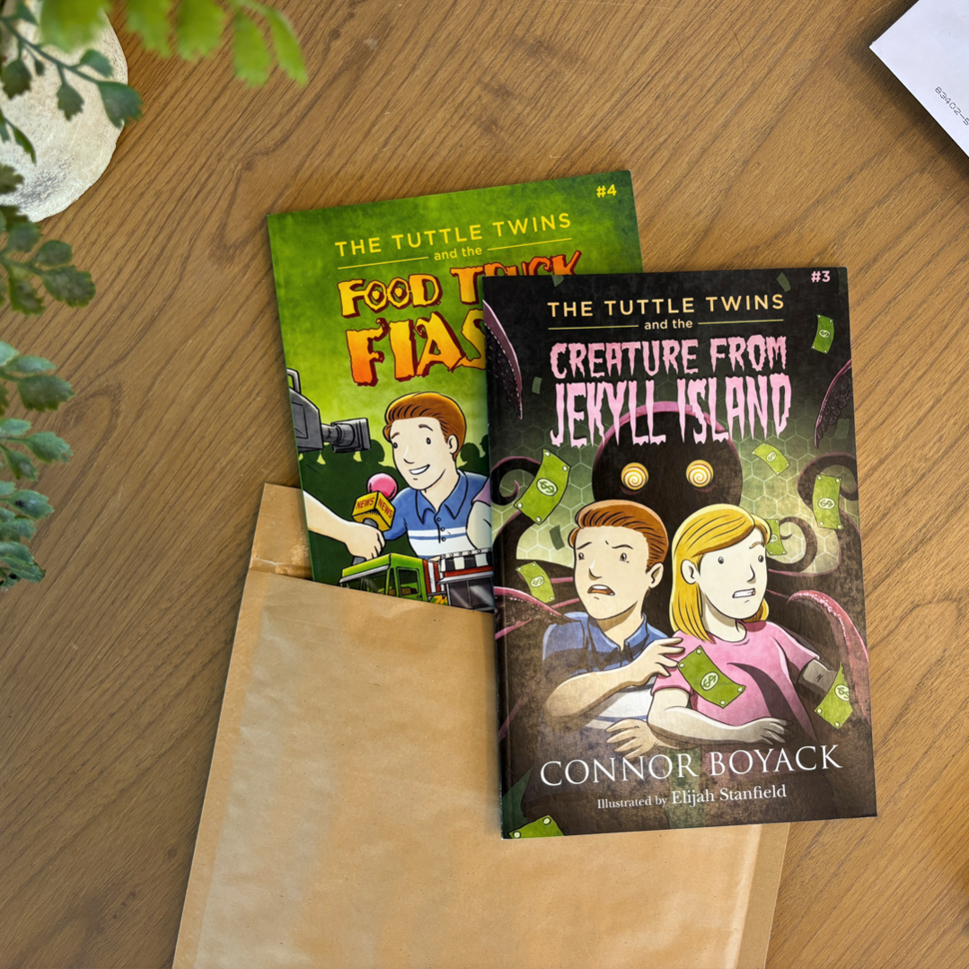 Two books titled 'Food Truck Fiasco' and 'Creature from Jekyll Island' by The Tuttle Twins on a wooden surface.