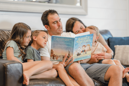 A father sitting on a couch, reading The Tuttle Twins: America’s History aloud to his wife and three children.