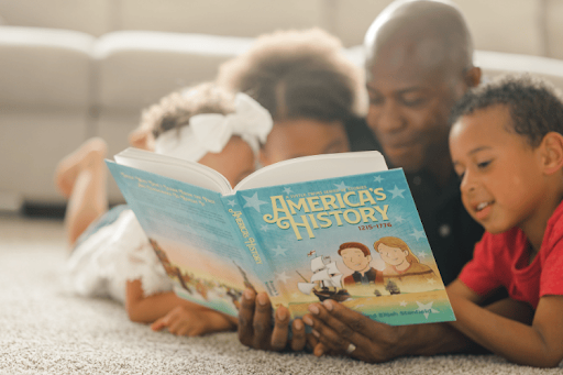 A man and three children lie on a carpeted floor while looking at an "America's History" book.