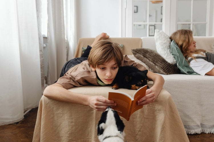 [Photo by Karola G on Canva.] A young boy lying comfortably on a beige sofa reading an orange book, accompanied by a small black dog resting beside him and a black-and-white dog looking up from the floor.