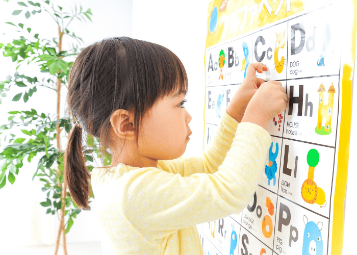 A young child reaches toward a colorful alphabet chart on the wall while identifying letters and pictures.