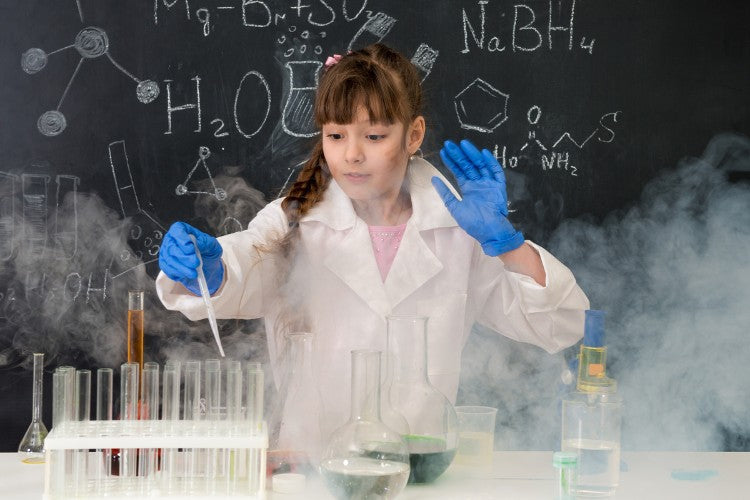 A child wearing gloves and a lab coat uses a dropper and test tubes while smoke rises during a chemistry experiment.