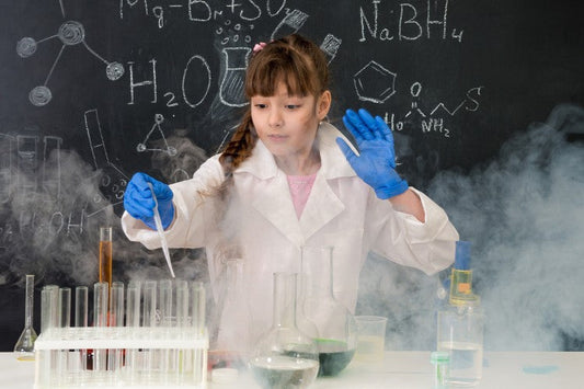 A child wearing gloves and a lab coat uses a dropper and test tubes while smoke rises during a chemistry experiment.