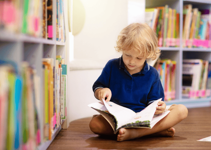 A young child sitting on the floor in a library, reading a picture book surrounded by shelves of books.