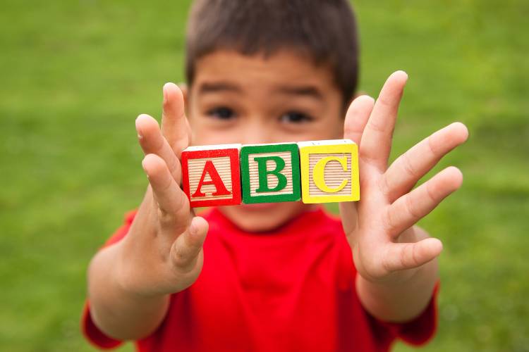Child holding up three alphabet blocks labeled A, B, and C in an outdoor setting.