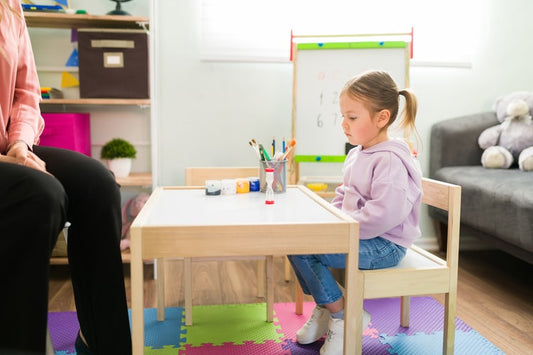 A young girl sitting quietly at a small table with art supplies, while an adult sits nearby.