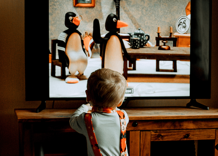 Rear view of a young child in a grey shirt and patterned suspenders standing in front of a television watching the Pingu animated show.