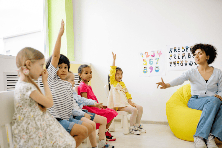 A diverse group of five young children sit facing a female teacher with curly hair, with two children raising their hands eagerly. Image by Yan Krukau on Canva