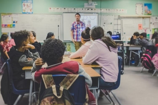 Students seated at desks listening to a teacher in a classroom setting.