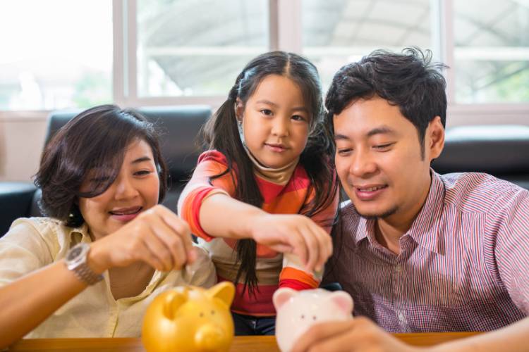 Parents and their daughter placing coins into piggy banks together at a table Photo Credit: Canva.