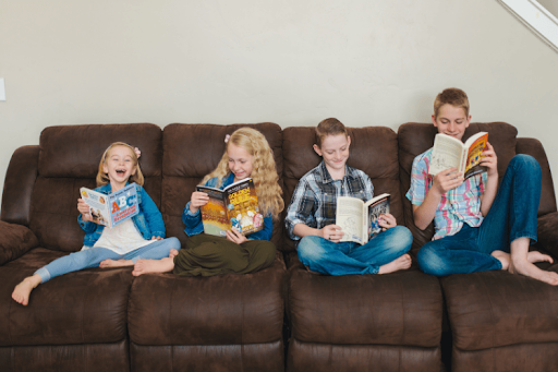 Four children sit side-by-side on a large brown leather sofa, each focused on reading their own Tuttle Twins book or magazine.