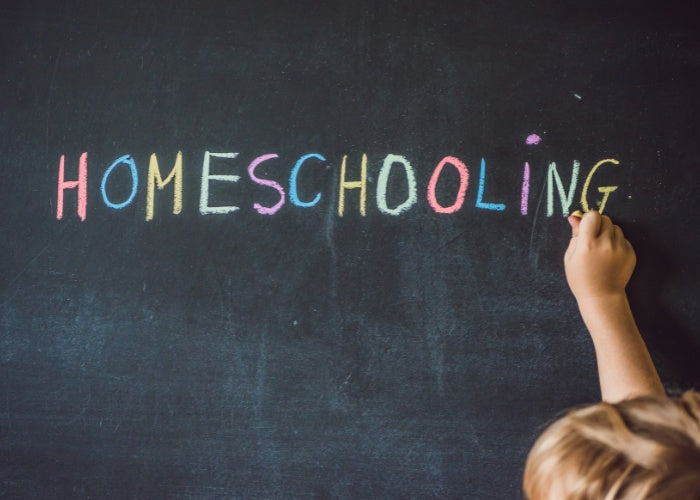 Child writing "HOMESCHOOLING" in colorful chalk on a blackboard.