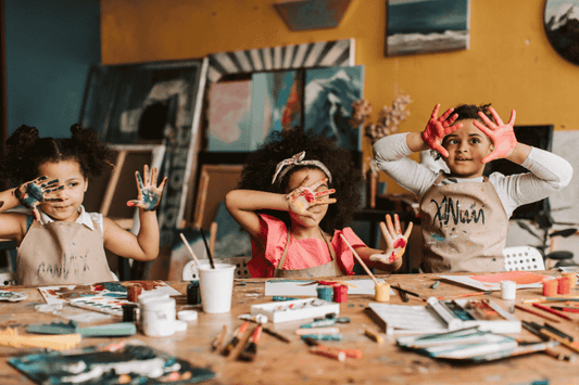 Three young girls wearing aprons in an art studio, smiling and holding up their hands which are covered in colorful paint. [Image by Vlada Karpovich on Canva.]