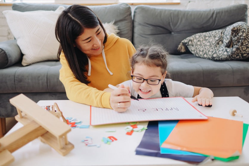 [Photo by Capturenow on Canva.] A smiling mother helping her young daughter with glasses practice writing the word apple on a small whiteboard, surrounded by colorful alphabet letters and educational wooden toys on a table.