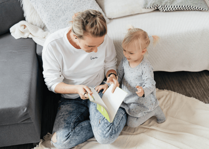 A parent sitting on the floor reading a book with a toddler in a cozy living room.