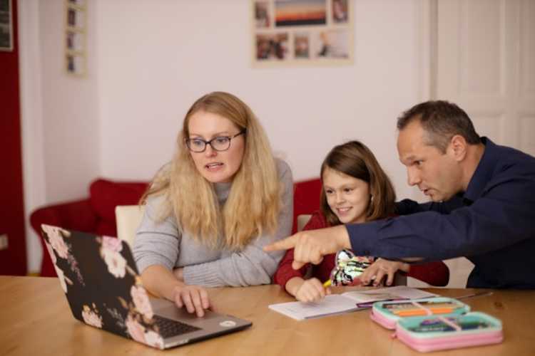 Two adults sit with a child at a table, pointing at a worksheet while using a laptop together.