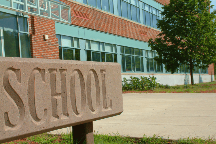A close-up of a concrete sign engraved with the word "SCHOOL," with a red brick school building visible in the background.