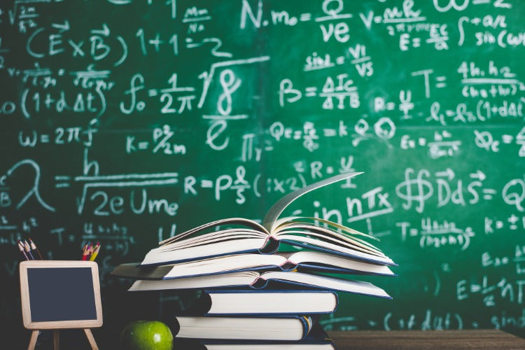 A stack of textbooks sitting on a desk in front of a green chalkboard filled with complex physics and math equations. [Photo by Patcharin from Canva.]