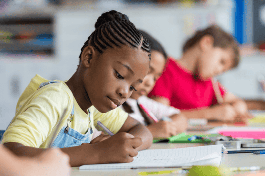 Young girl writing in a notebook at her classroom desk with other students working in the background.
