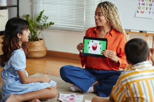 A teacher shows an educational spelling game on a tablet to two children sitting on a rug.