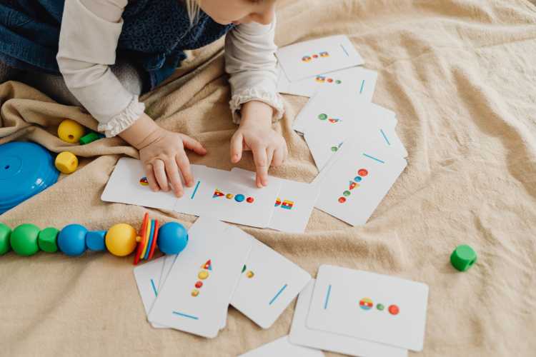 A toddler sits on a blanket and points at counting cards while sorting colorful wooden beads.