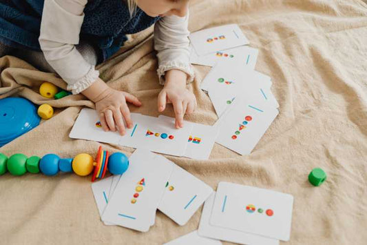 A toddler sits on a blanket and points at counting cards while sorting colorful wooden beads.