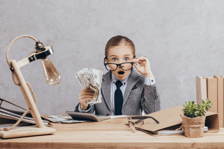A young girl dressed in a grey business suit and glasses looking shocked while holding a handful of cash at a desk.