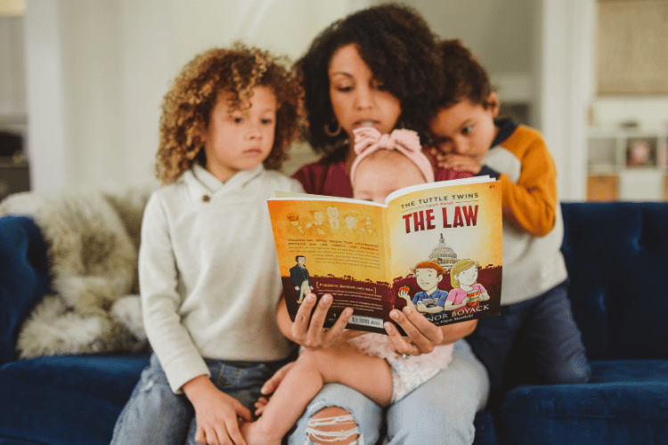 A mother sitting on a blue couch reading The Tuttle Twins Learn About The Law aloud to her three children.