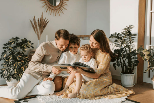 A family of four sits closely together on a white rug, focused on reading a large book in a sunlit room.