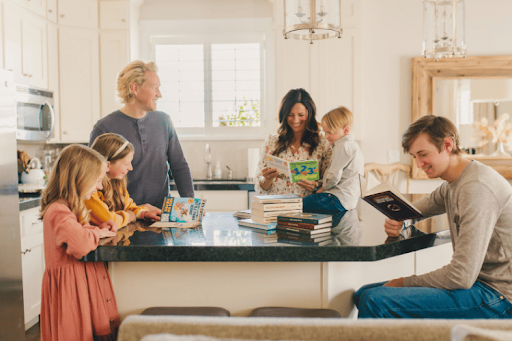 A family of six gathers around a kitchen island, each person engaged in reading different books.