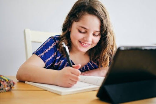 Homeschooled girl smiling while writing notes during an online lesson.