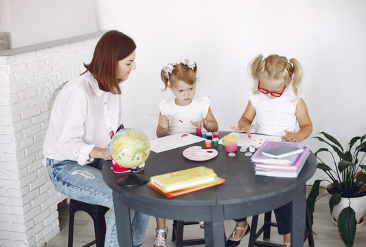 A teacher sitting with two young girls at a table, guiding them as they paint with colorful art supplies.