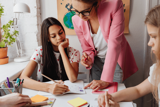 A teacher in a pink blazer assists a student with her notebook in a classroom.