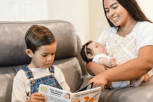 Young boy reading toddler book while mother holds baby.