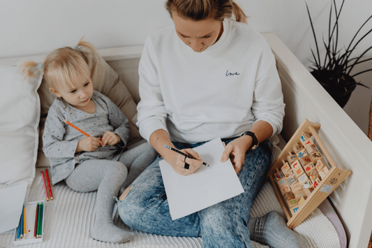 A woman draws on a piece of paper while a toddler girl watches closely, sitting together on a white daybed with educational toys nearby.