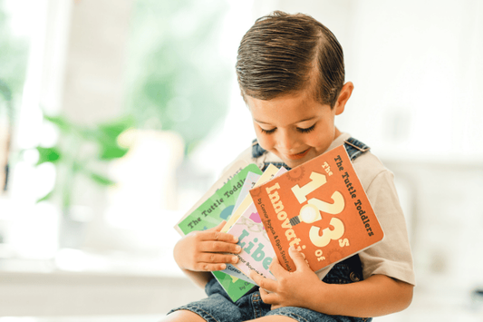 A young boy in denim overalls smiling down at a fan of "The Tuttle Toddlers" board books he is holding.