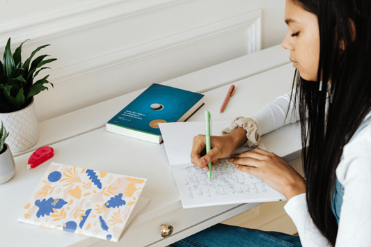 A young lady writes in a notebook at a clean white desk next to a potted plant and books.